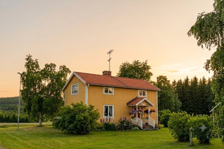Yellow house with red roof, surrounded by trees and grass at sunset.
