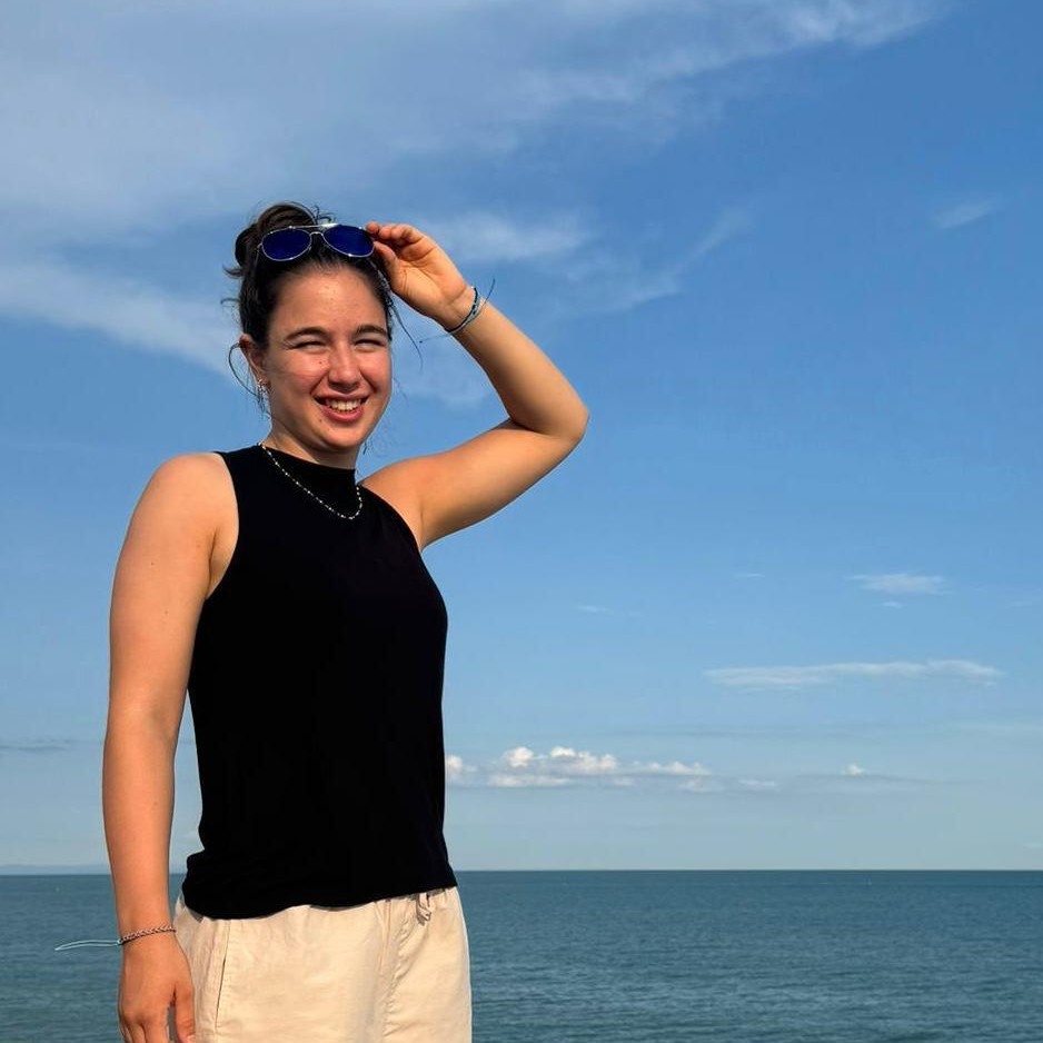 Person in black top by the sea, smiling and holding sunglasses, with a clear blue sky in the background.
